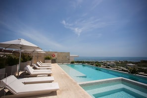 Piscina no terraço com espreguiçadeiras e guarda-sóis, com vista para a costa.