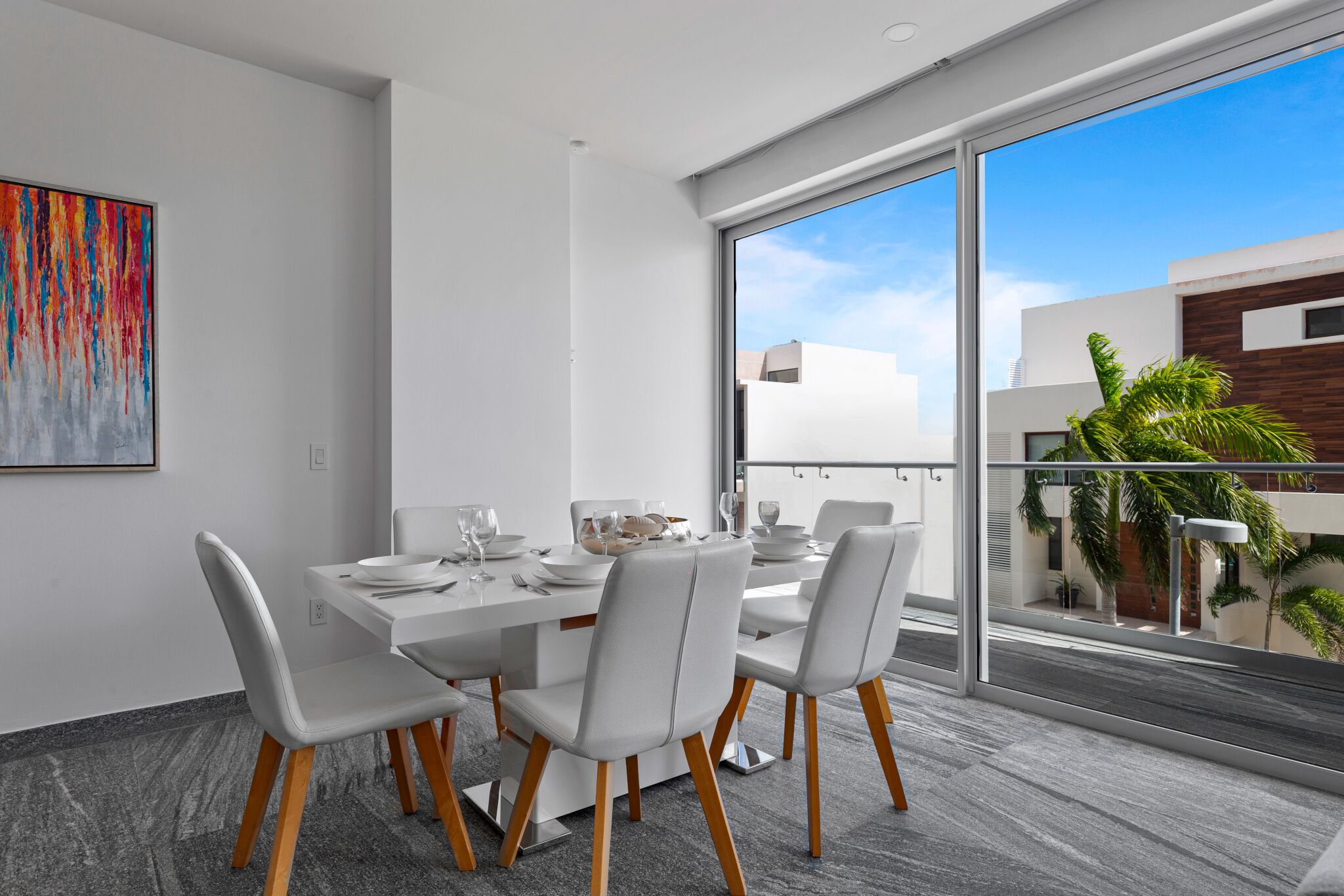 A modern dining area with a white table set for four, large windows offering a view of the cityscape, and a colorful abstract painting on the wall.