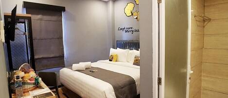 Una habitación de hotel con una cama, una mesita con una cesta de frutas, una pared con un mensaje alegre y un espacio con ducha.