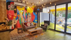 A modern hotel lobby with a colorful abstract mural, a reception desk, comfortable seating, and large windows offering a view of the outside. - Nine Hotel Palembang (Palembang)