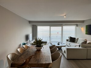 Una sala de estar moderna con una mesa de comedor de madera, sofá color beige y ventanas grandes con persianas.