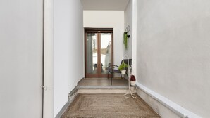 Un corridoio stretto con una porta in legno alla fine, un piccolo tavolo con sedie e una pianta in vaso su un supporto.