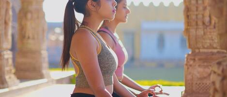 Dos personas sentadas sobre esterillas de yoga en un espacio al aire libre con columnas arquitectónicas.