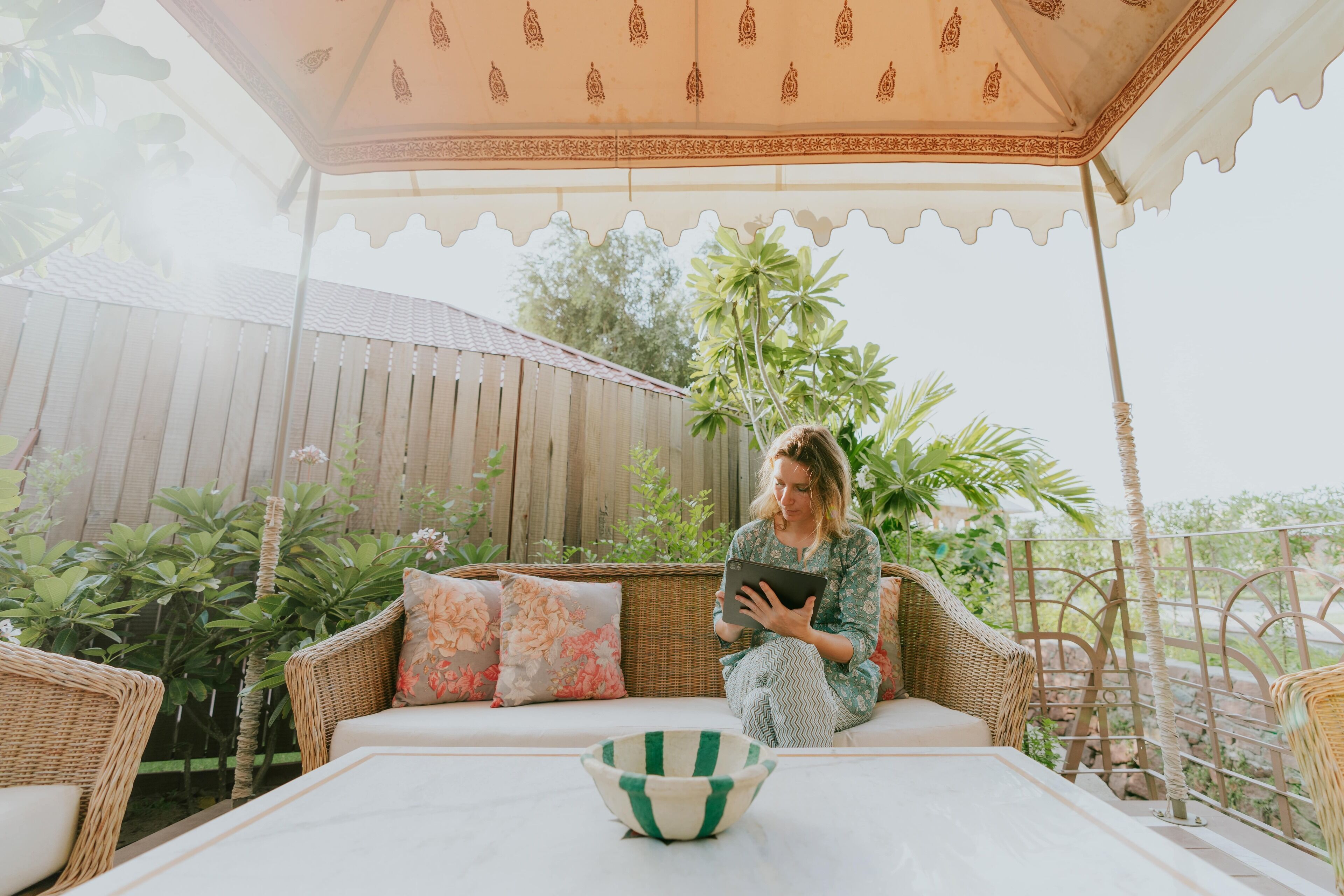 Una persona sentada en un sofá de mimbre bajo un dosel estampado, sosteniendo una tableta.