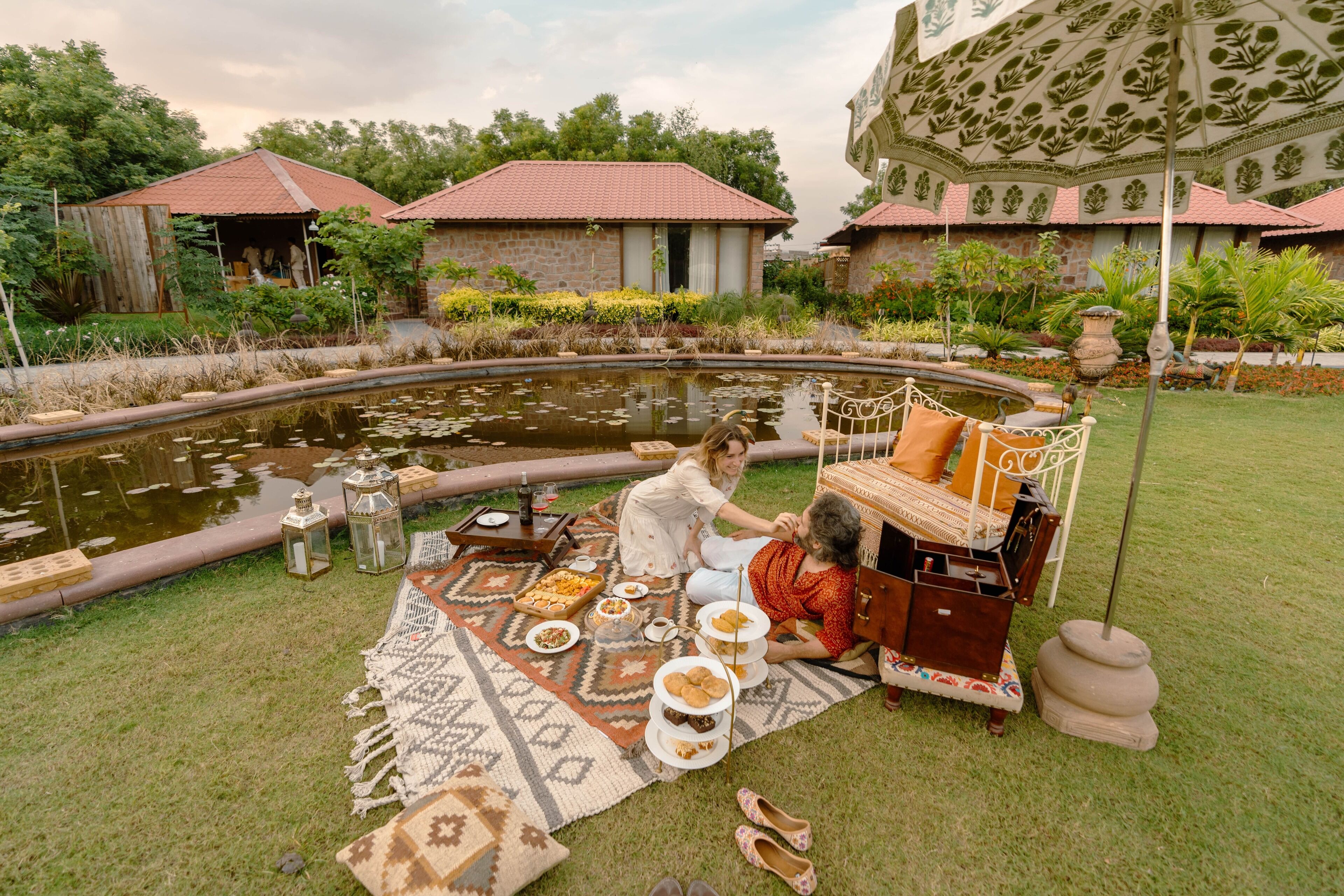 Egy piknikhez előkészített helyszín: étkezőasztal, székek és párnákkal ellátott pad található itt. A háttérben egy kis tó, melyen vízililiomok úsznak, és egy pavilon is látható.