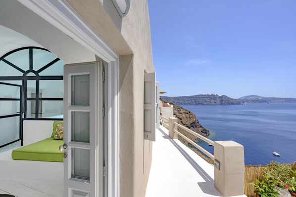 A balcony with a view of the sea, a door to a room with a green bed, and a white exterior wall. - Nano Oia Canaves (Santorini)
