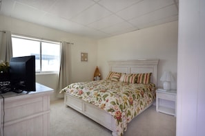 Une chambre à coucher avec un lit, une table de chevet avec une lampe, une commode, un téléviseur posé sur la commode et une fenêtre avec des rideaux.