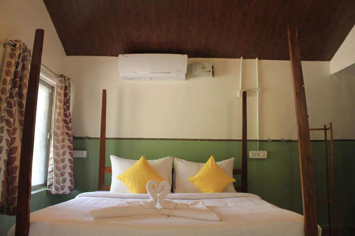 A hotel room with a bed, two pillows, a towel, a window with curtains, an air conditioning unit, and a wooden ladder.