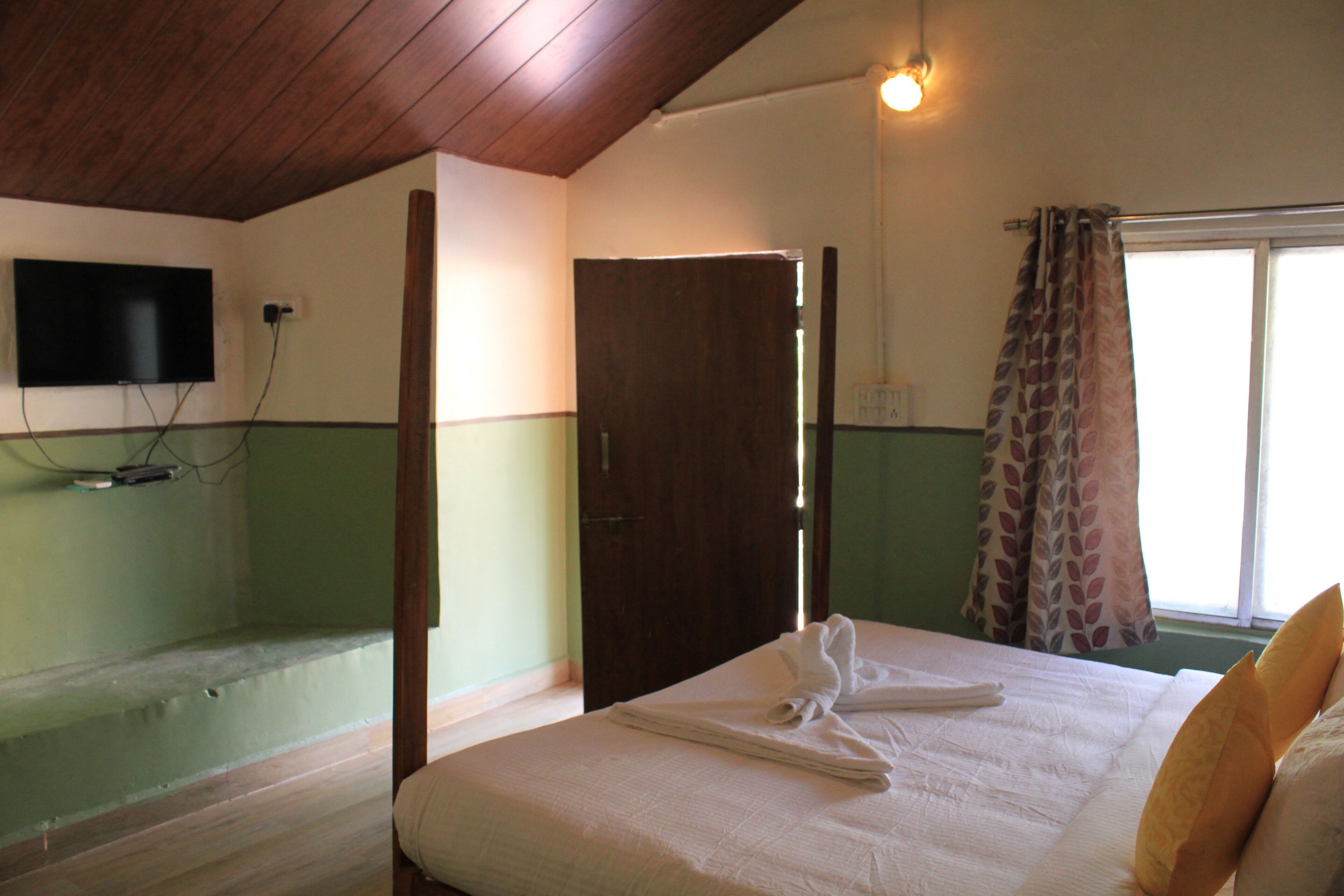 A hotel room with a bed, a television mounted on the wall, a window with curtains, and a wooden ceiling.