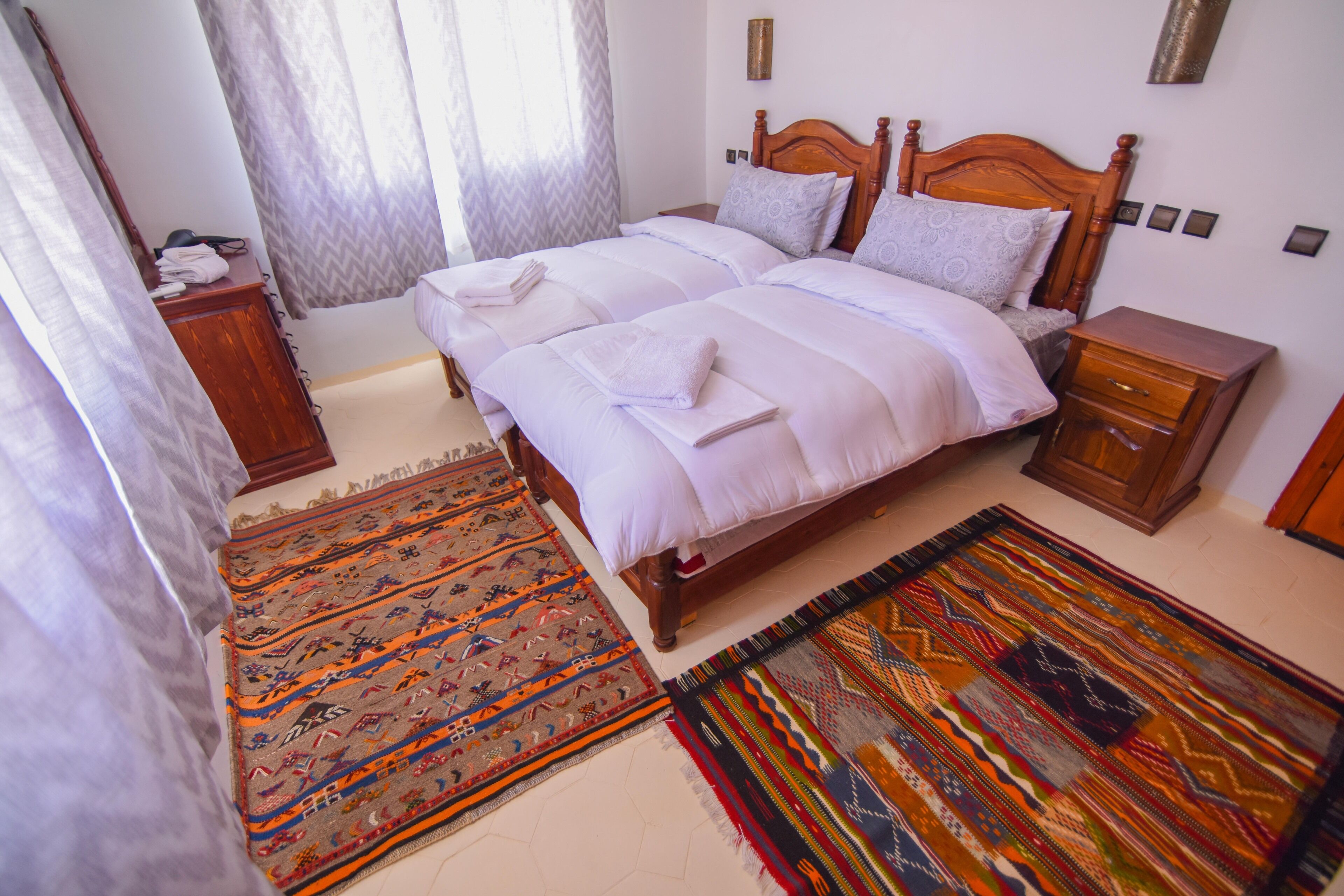 A double bed with white linens, a wooden headboard, and a nightstand with a drawer. There are two patterned rugs on the floor.