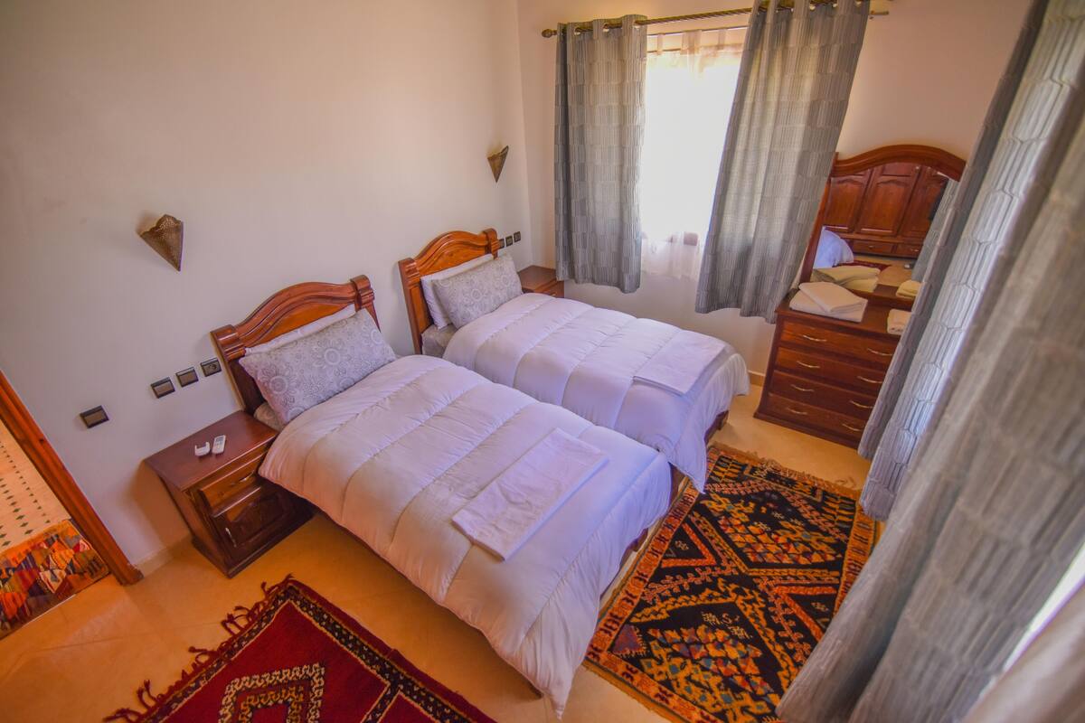Une chambre avec deux lits, une commode en bois et un tapis à motifs.