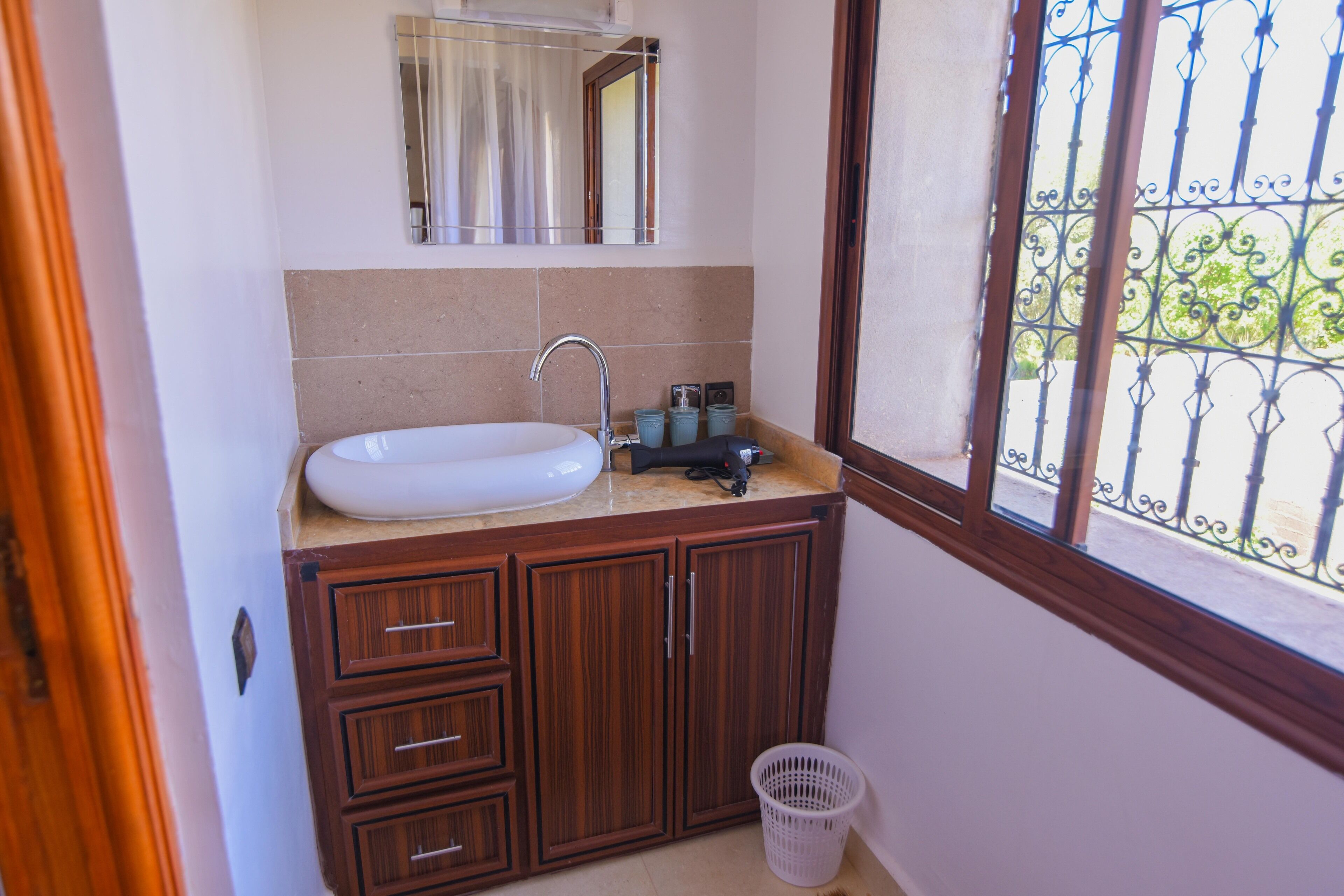 Un baño con lavamanos blanco, armarios de madera, un espejo y una ventana con reja de metal.