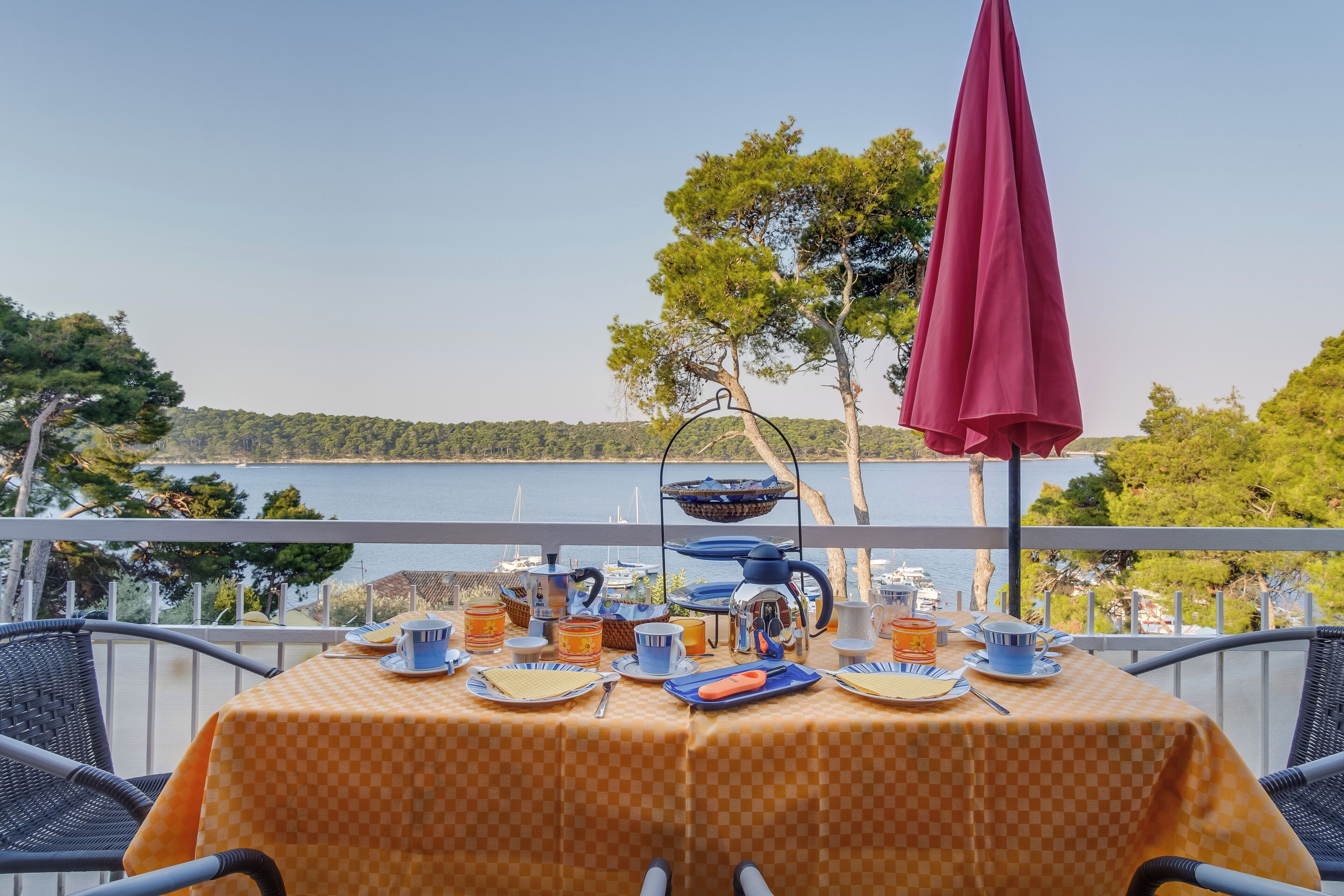 Una mesa lista para el desayuno con vista al mar, un paraguas rojo y asientos al aire libre.