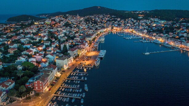 Vista aérea de un pueblo costero al anochecer, con un puerto deportivo lleno de barcos, una imponente aguja de iglesia y una zona urbana densamente poblada con edificios iluminados.