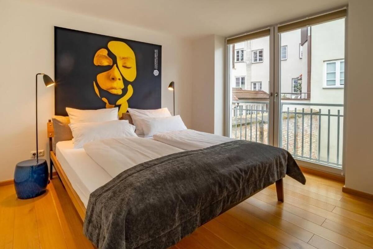 A bedroom with a large bed, a black and yellow artwork on the wall, and a balcony with a view of buildings.