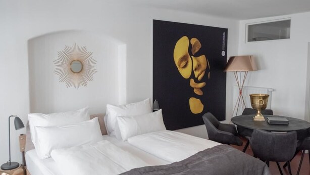 A modern hotel room with a large bed, a round table with chairs, a lamp, and a wall-mounted decorative piece. - ANA Suites Augsburg City Center (Augsburg)