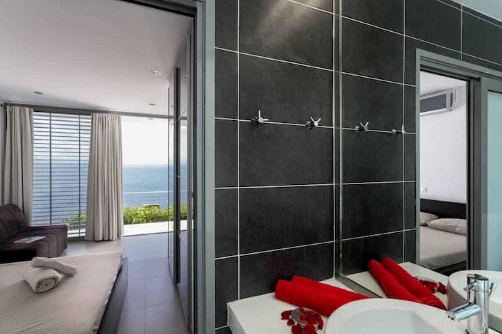A modern bathroom with a large mirror, dark tiled walls, and a view of the sea.