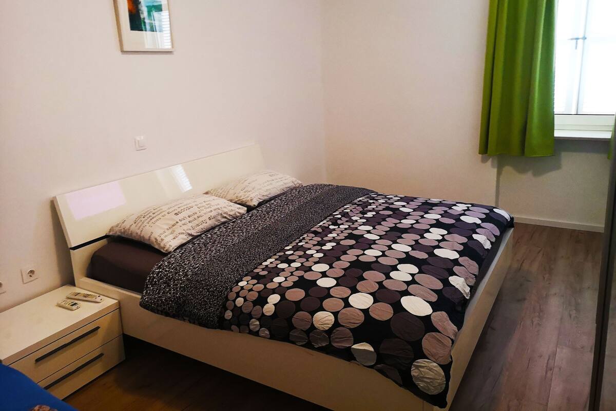 Ett sovrum med en säng, ett nattduksbord, en låda, en inramad tavla på väggen och ett fönster med gröna gardiner.