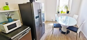 A compact kitchen with a refrigerator, microwave, and dishwasher. There's a round table with chairs and a window with plants. - Chillax Rooms (Split)