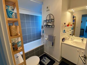 A bathroom with a bathtub, toilet, and sink. There is a shelf with toiletries and a towel rack.