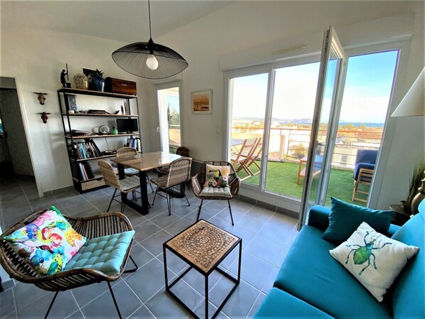 A spacious living area with a dining table, wicker chairs, a blue sofa, and a large window offering a view of the outdoors.