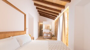 A modern hotel room with a large bed, wooden ceiling beams, a bathroom with a mirror, and a patterned throw on the bed. - Petit Palau Alcudia Boutique Hotel (Alcúdia)