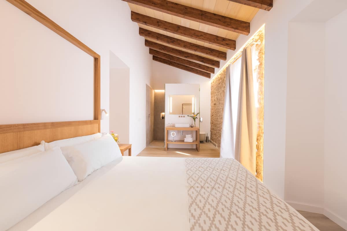 A modern hotel room with a large bed, wooden ceiling beams, a bathroom with a mirror, and a patterned throw on the bed.