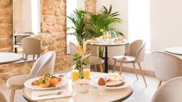 A breakfast setting with a stone wall, a potted plant, and a table with plates of food, glasses of orange juice, and a bowl of cereal.