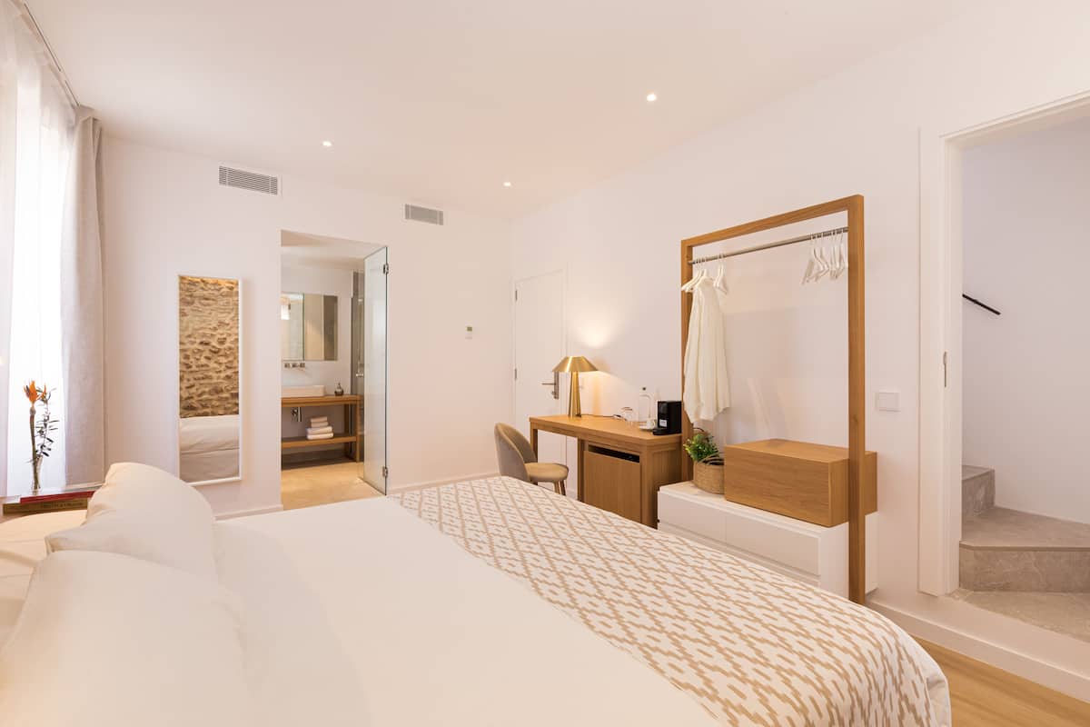 A modern hotel room with a large bed, a desk, a mirror, and a bathroom visible through an open door.