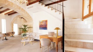A spacious living area with a wooden desk, a sofa, and a dining table. There is a staircase with a wooden railing and a painting on the wall. - Petit Palau Alcudia Boutique Hotel (Alcúdia)