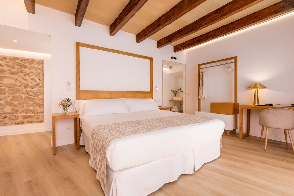 A bedroom with a large bed, wooden beams, a stone wall, a desk, and a mirror.