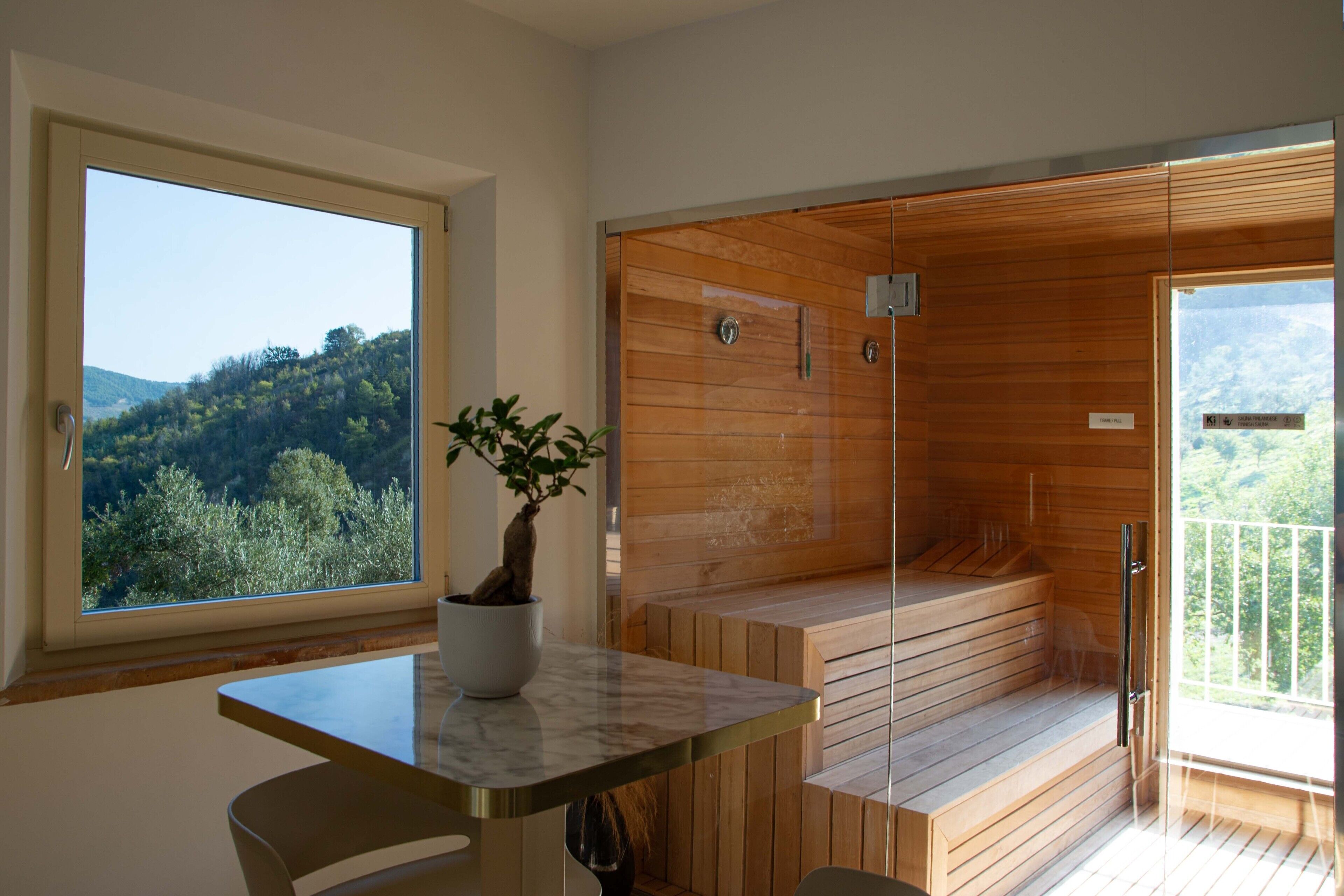 Un baño moderno con sauna de vidrio, vista al bosque y una mesa de mármol con planta en maceta.