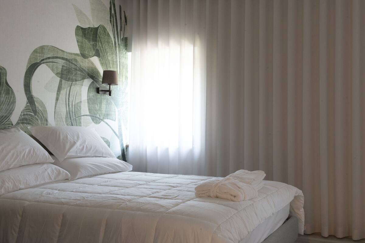 A neatly made bed with white linens and a folded towel, a wall-mounted lamp, and sheer curtains allowing natural light.