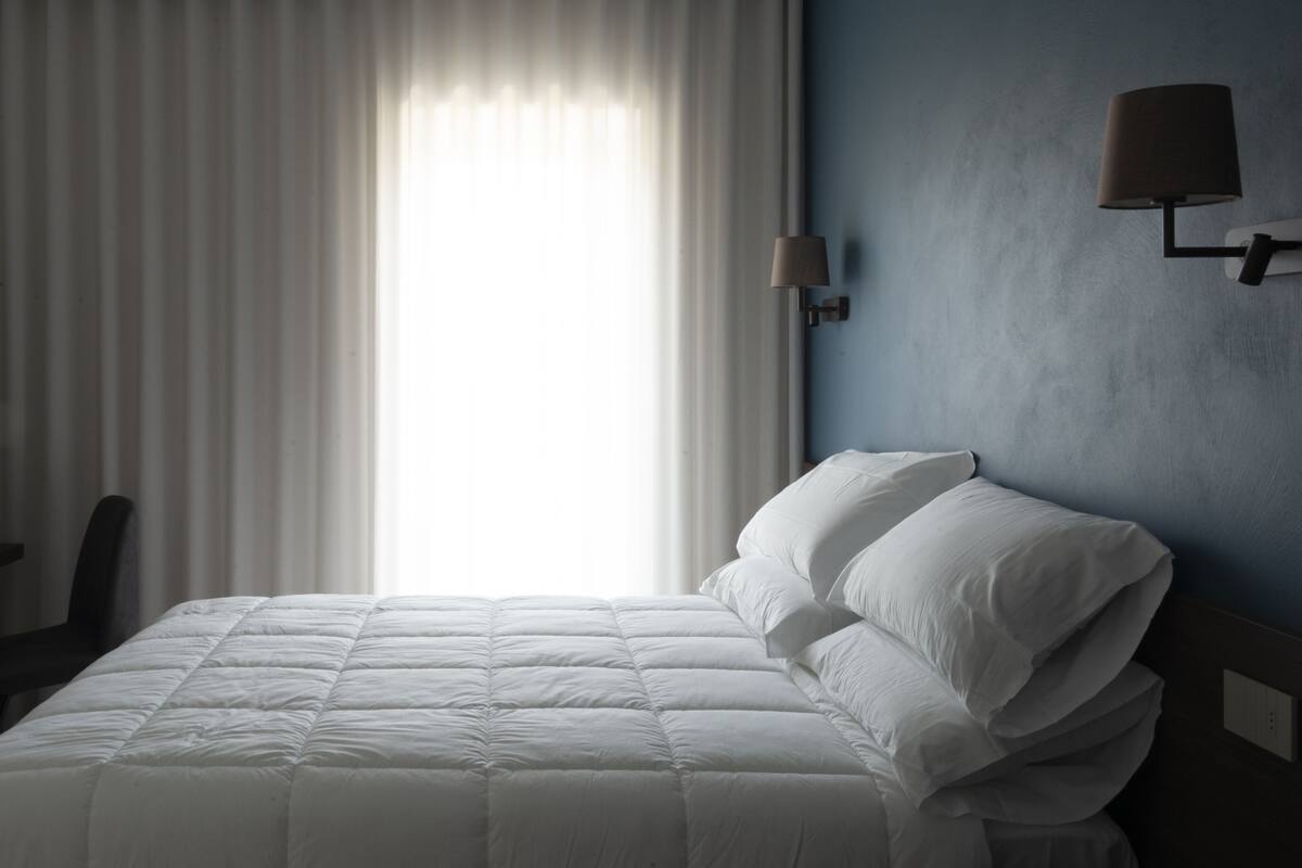 A hotel room with a bed, a desk, a chair, a lamp, and a window with curtains.