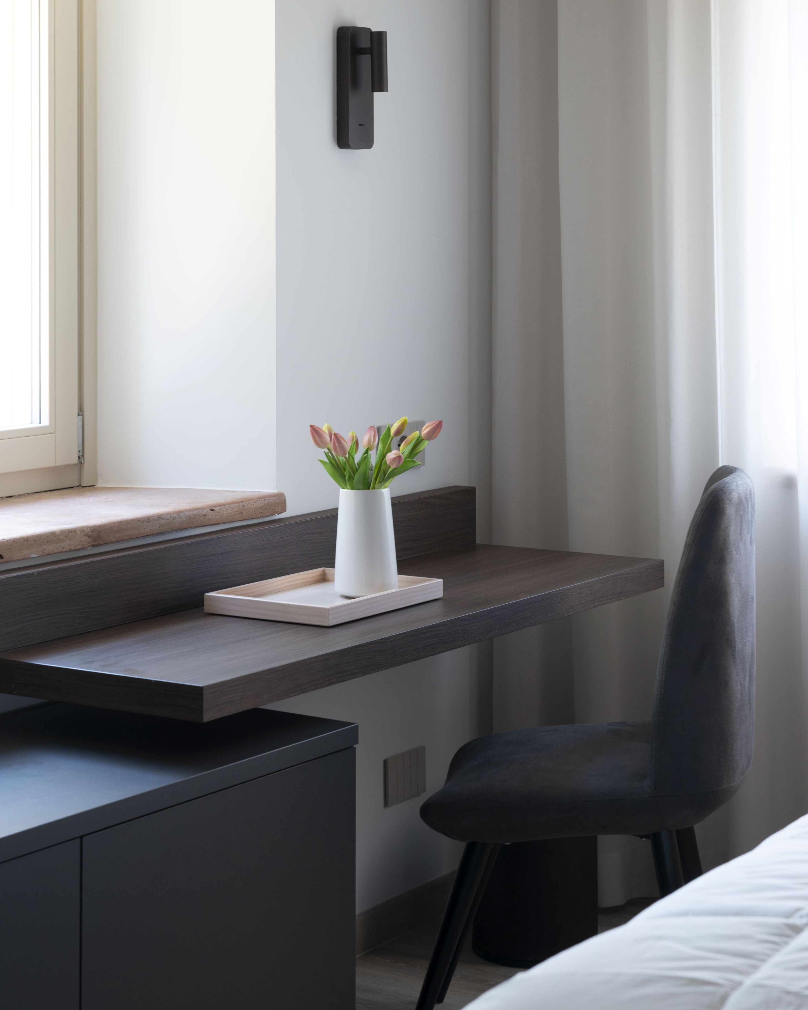 Una habitación de hotel moderna con un escritorio de madera, un jarrón blanco con flores, una silla negra y una cama con ropa de cama blanca.