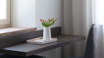 Una camera d'albergo moderna con una scrivania in legno, un vaso bianco con fiori, una sedia nera e un letto con biancheria da letto bianca.