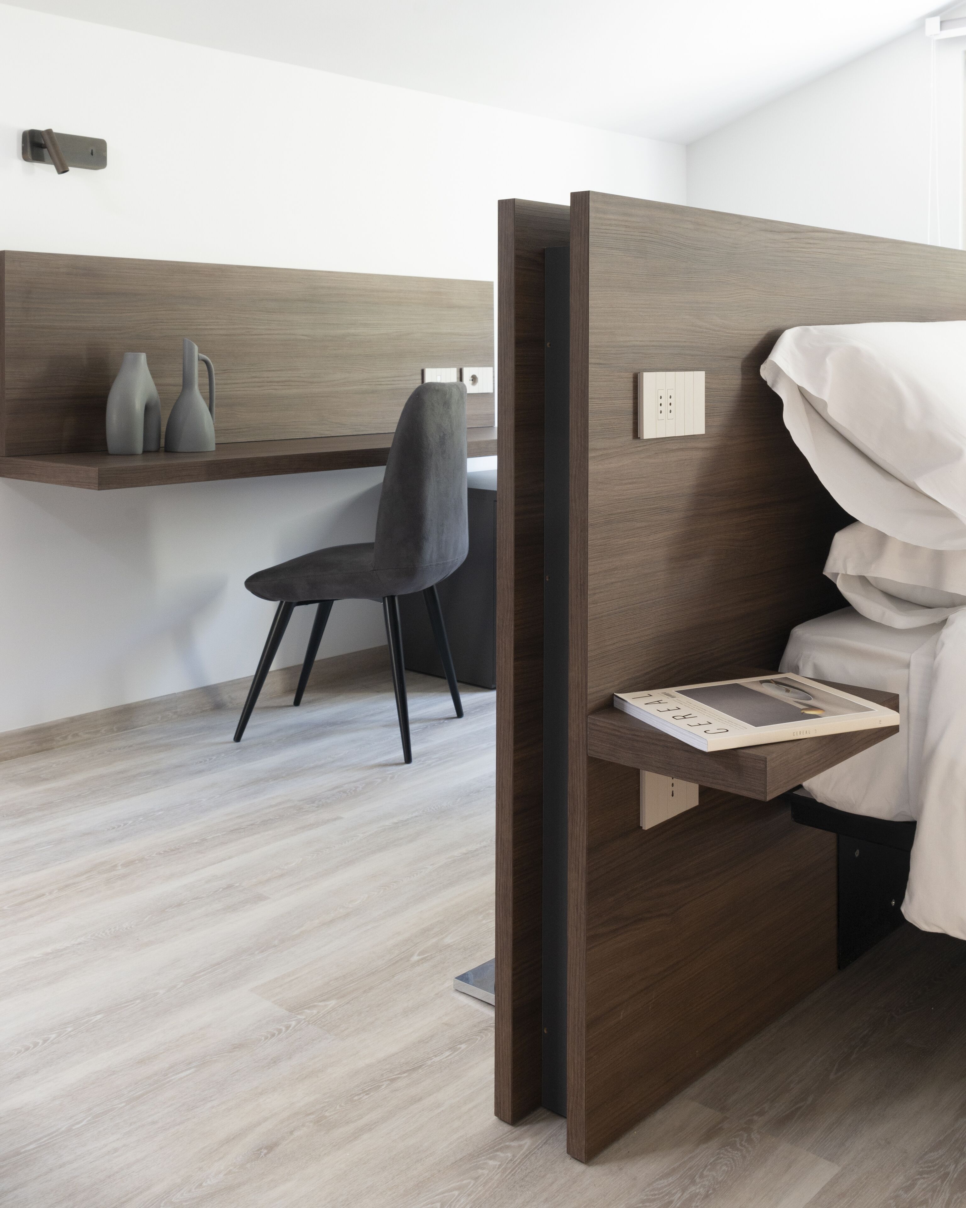 Un dormitorio moderno con cabecera de madera, una mesita de noche con un libro, una silla y un estante con objetos decorativos.