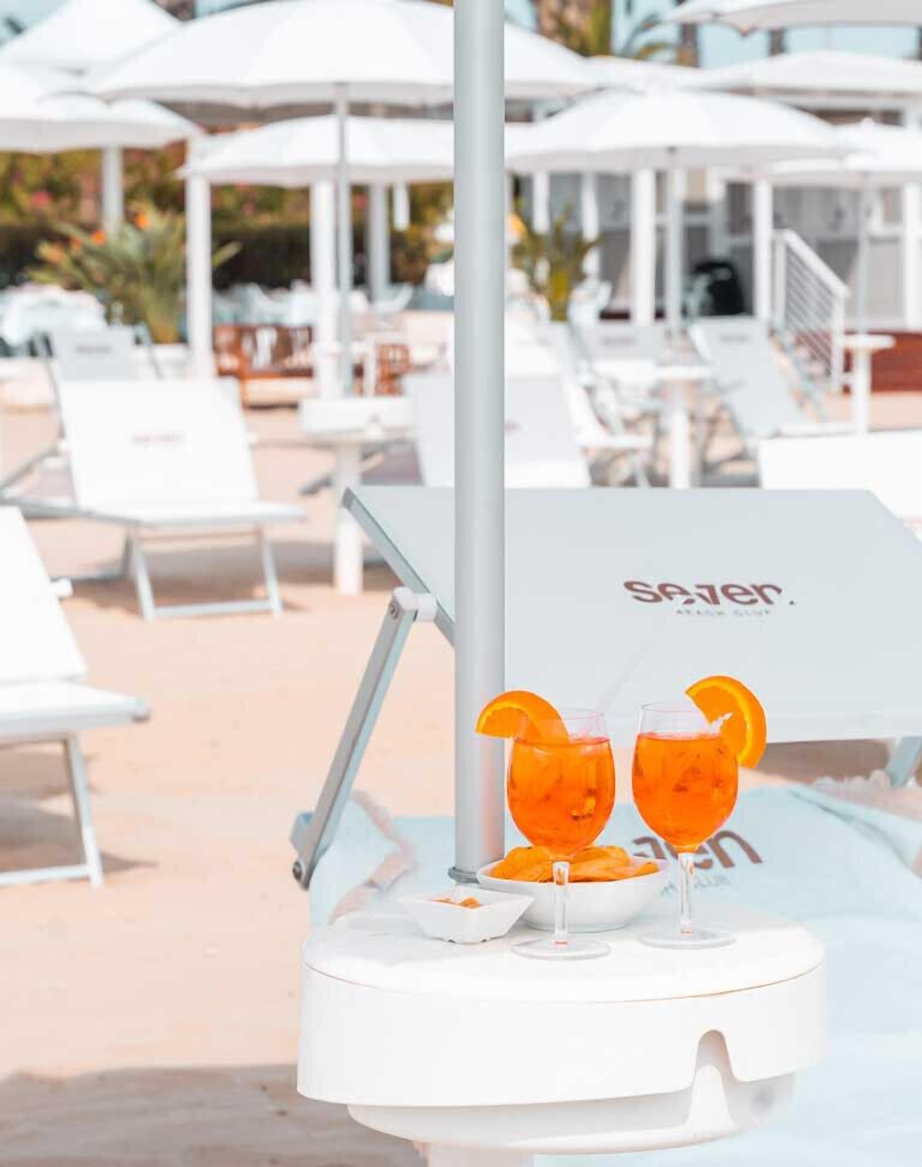 Area piscina con lettini e ombrelloni, distributore di bevande con due bicchieri di arancia e un cartello con su scritto "Sazer".