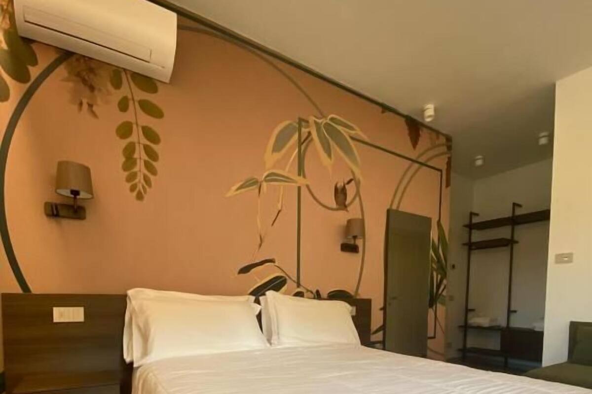 A hotel room with a large bed, a wall-mounted air conditioner, a wooden nightstand, a lamp, and a plant.