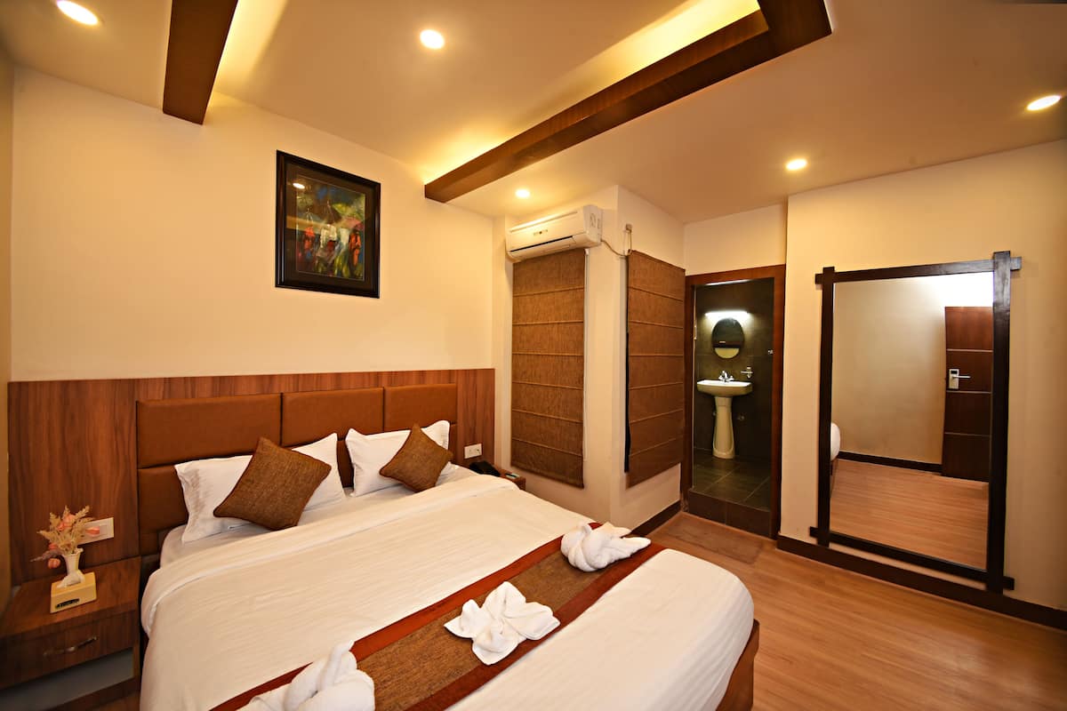 A hotel room with a large bed, wooden headboard, a painting on the wall, an air conditioning unit, a bathroom with a sink and mirror, and wooden flooring.