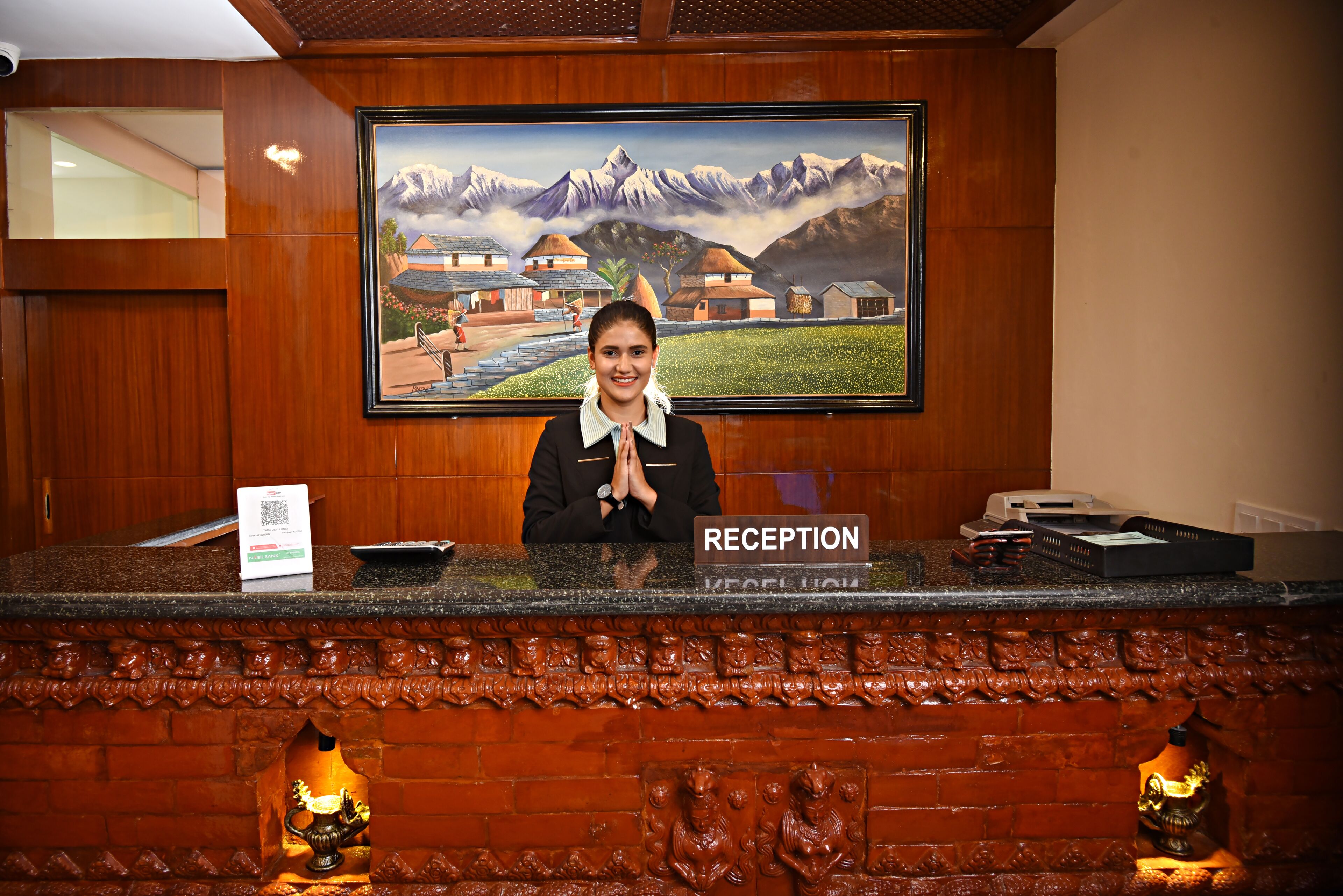 A reception desk with a "RECEPTION" sign, a framed painting of a mountainous landscape, and a wooden reception area with carved details.