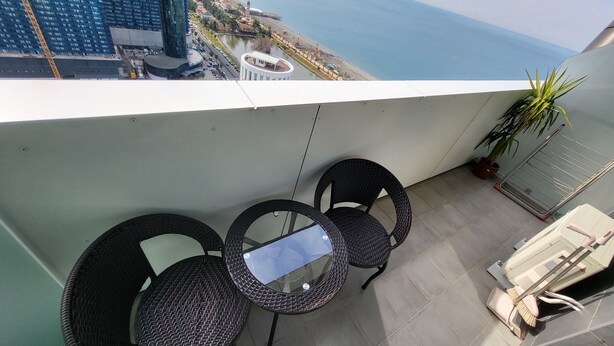 A balcony with two wicker chairs and a small table, overlooking a coastal area with buildings and a body of water. - Black Sea Apartments Alliance Palace (Batumi)