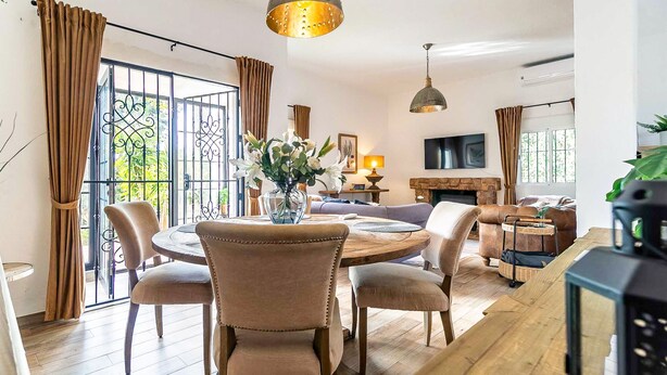Een ruime woonkamer met een ronde houten eettafel, zithoeken, een open haard en grote ramen met gordijnen.