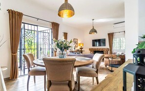 Amplia sala de estar con una mesa de comedor redonda de madera, asientos cómodos, chimenea y ventanas grandes con cortinas.