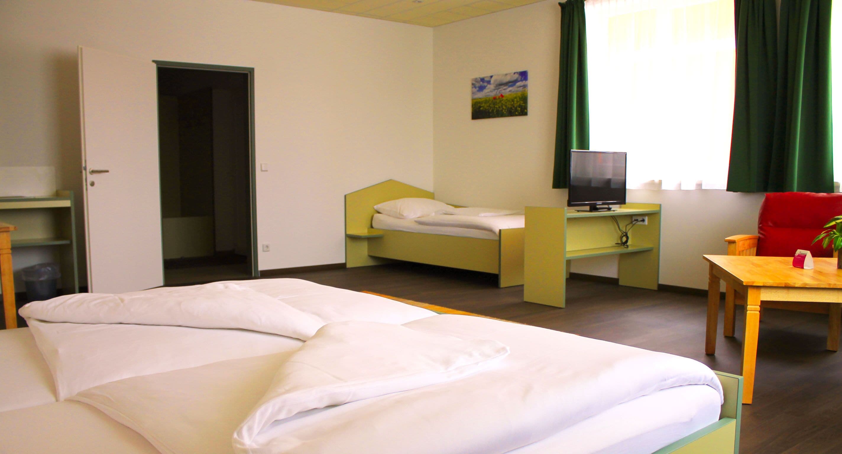 Habitación de hotel con cama, escritorio, computadora, silla y ventana con cortinas.