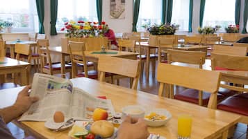A dining area with wooden tables and chairs, a person reading a newspaper, and a breakfast spread including fruits, bread, and juice.