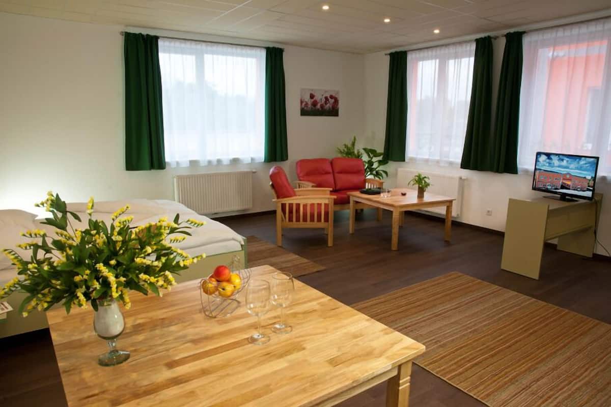 A well-lit room with a bed, a wooden table with fruits and glasses, a red sofa, a small table with a plant, and a TV on a stand.