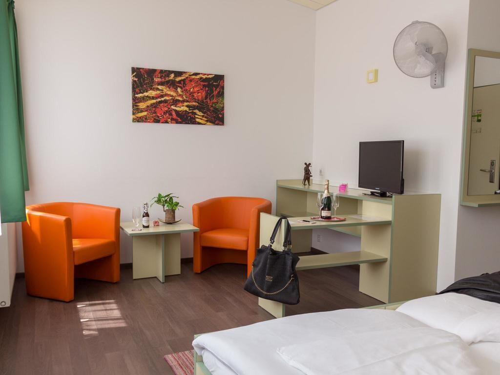 A hotel room with a bed, two orange chairs, a desk with a TV, a painting on the wall, and a fan.