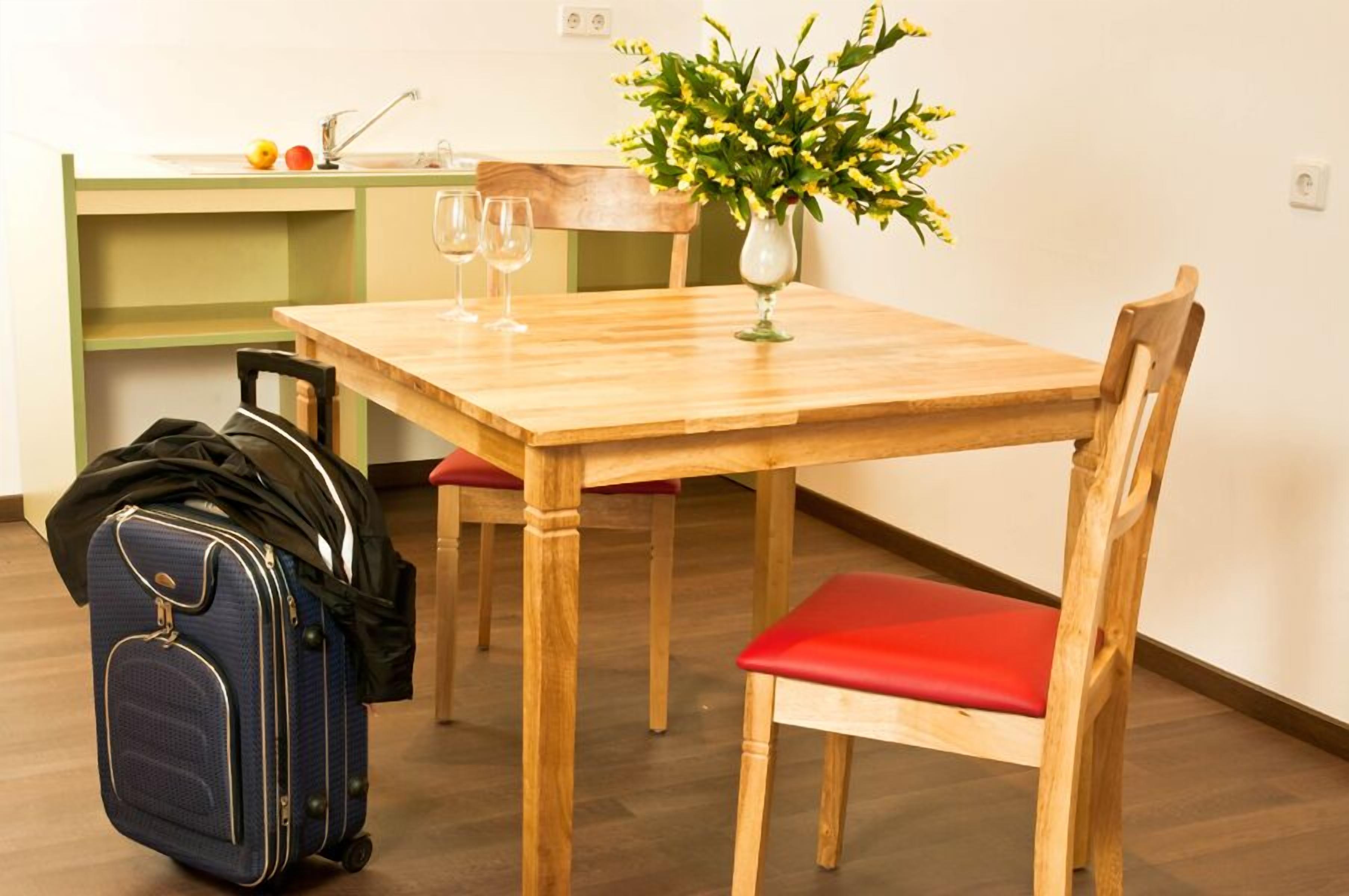 A dining area with a wooden table, red cushioned chairs, a vase with yellow flowers, and a blue suitcase on the floor.