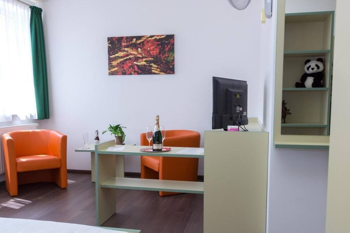 A hotel room with a bed, two orange chairs, a small table with a bottle and glass, a TV, and a shelf with a panda plush toy.